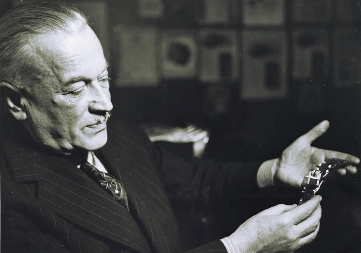Black-and-white photo: an older man in a pinstripe suit looks at a wristwatch in his hand