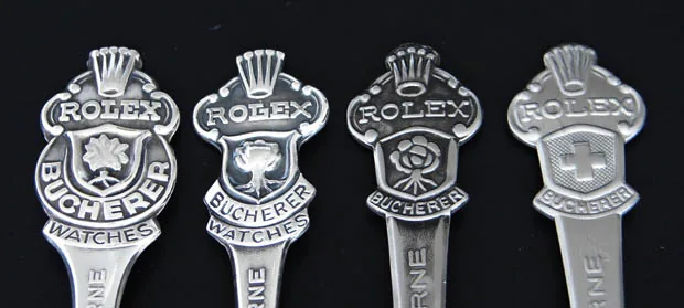 Top ends of four Rolex spoons with crown, Rolex lettering, Bucherer shield and various coats of arms on a black background