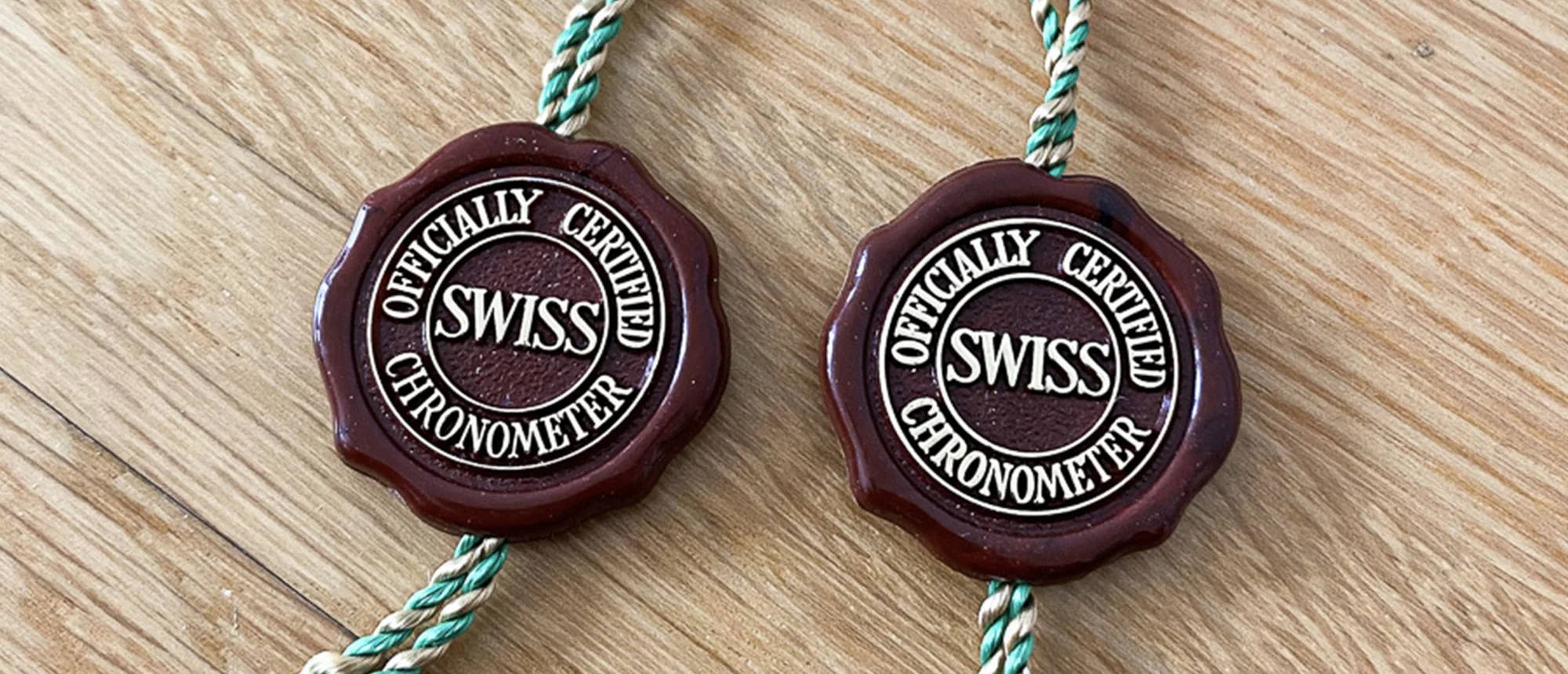 Zwei braune Chronometer-Siegel mit der Aufschrift „Officially Certified Swiss Chronometer“ an grün-beigen Kordeln auf einem Holztisch